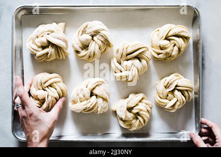 Bäcker macht Kardamom-Brötchen. Helles und weißes Bild von oben, in dem Bäckerhände ungekochte schwedische Kardamombrötchen auf ein zum Backen bereitetes Tablett legen. Stockfoto