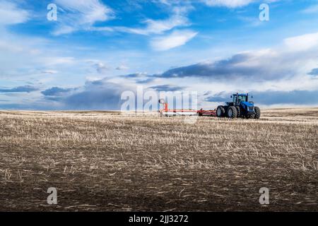 Ein achträdriger Traktor, der einen Pflug über ein bebautes Feld mit sanften Hügeln in Rocky View County Alberta Canada unter einem dramatisch bewölkten Himmel zieht. Stockfoto