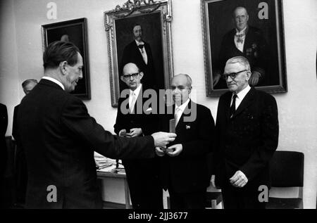 Örebro Skofabrik schließt, der bezirksrat teilt Auszeichnung, Weihnachtsbäume 19. Dezember 1967 Herr in schwarzem Anzug steht mit dem Rücken gegen die Kamera und hält ein Ding in seiner rechten Hand, das er ausgestreckt hält. Im Hintergrund sind drei Herren in schwarzen Anzügen, weißen Hemden und hellen Krawatten zu sehen. Zwei von ihnen halten kleine Schmuckkisten in ihren Händen. An der Wand hinter ihnen hängen drei Gemälde. Stockfoto