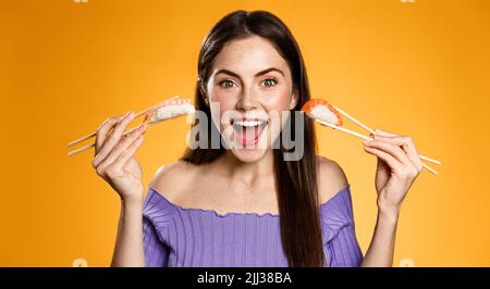 Lächelnde Frau, die Sushi zeigt, Nigiri mit Essstäbchen hält, aufgeregt aussieht, Werbung für asiatisches Restaurant oder Essen zum Mitnehmen, orange Stockfoto