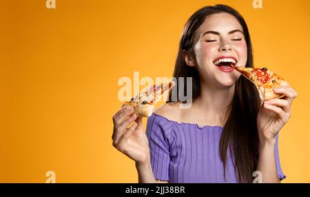 Hungrige lächelnde Frau nimmt einen Bissen Fast Food, hält zwei Pizzascheiben, isst mit Vergnügen, oranger Hintergrund Stockfoto