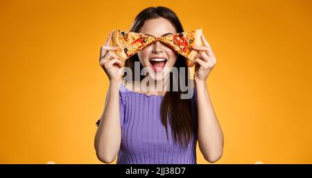 Fröhliche lächelnde Frau, die Pizzablecken auf den Augen mit aufgeregter Gesichtsausdruck hält, Konzept der Pizzalieferung, Essen zum Mitnehmen und Fast Food essen Stockfoto