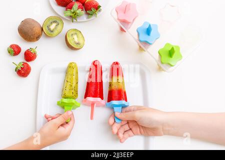 Zwei Hände für Kinder, nehmen Sie mehrfarbige hausgemachte Kiwi- und Erdbeereis von einem weißen quadratischen Teller Stockfoto