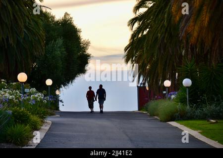 Zwei Liebhaber machen einen Spaziergang bei Sonnenuntergang auf einer ruhigen Straße oder Gasse mit Straßenlaternen und Palmen, die sie in Rutherglen umrahmen Stockfoto