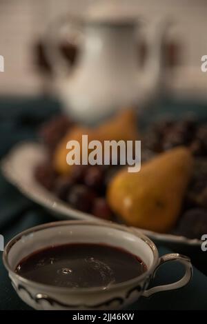 Der tiefrote Tee in einer weiß-silbernen china-Tasse sitzt vor einer Platte aus violetten Muscadinen und Bosc-Birnen, mit einer passenden Teedose im Hintergrund Stockfoto
