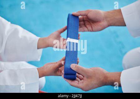 Heute ein blauer Gürtel, morgen ein schwarzer Gürtel. Ein unverkennbarer Mann, der seinem jungen Studenten einen blauen Karate-Gürtel in einem Studio überreicht. Stockfoto