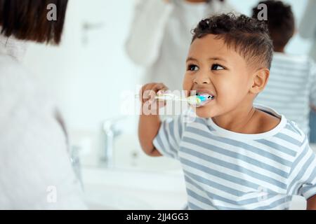 Schaut Mama, ich habe jeden Zahn. Ein entzückender kleiner Junge putzt sich die Zähne, während seine Mutter ihm zu Hause hilft. Stockfoto