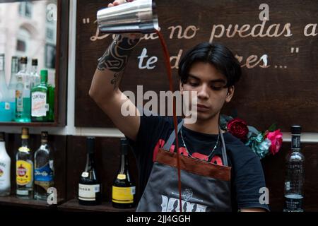 Ein Latino-Barkeeper, der in einem Restaurant arbeitet. Der Barkeeper bereitet ein Getränk mit einem Flüssigkeitsstrahl in einem Shaker zu. Ein Barkeeper in einer Theke. Es gibt viele Alkoholflaschen im Hintergrund. Stockfoto