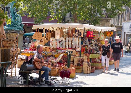 Limoux Aude France 07.22.22 Sommermarktplatz Szene. Männlicher Darsteller singt und spielt Gitarre. Stall mit bunten Taschen und Sonnenhüten. Reifer Mann A Stockfoto