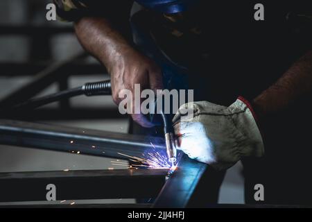 Der Handwerker, der an seinem Arbeitsplatz in der Werkstatt Schweißen und Schleifen durchführt, während die Funken um ihn herum „fliegen“. Er trägt einen Schutzhelm und Stockfoto