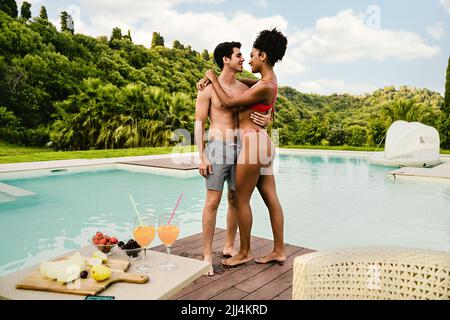 Romantische junge Freund und Freundin stehen während des Sommerurlaubs am Pool und umarmen sich - auf dem Beistelltisch Obst, Getränke und gesunde tr Stockfoto