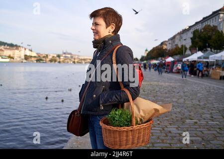 Frau auf dem Bauernmarkt am Ufer der Moldau in Prag lächelt und hält einen Korbkorb mit frischen Mikrogrüns in der Hand Stockfoto