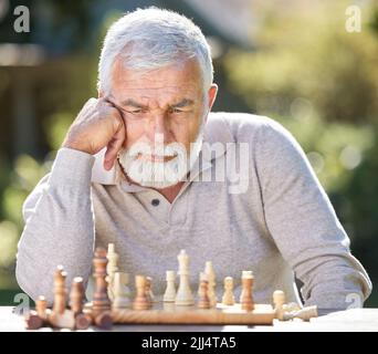 Wo soll ich mich als nächstes bewegen. Ein älterer Mann, der allein draußen sitzt und nachdenklich schaut, während er ein Schachspiel spielt. Stockfoto