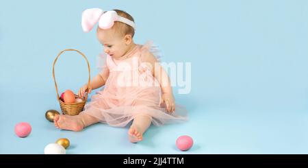 Happy Baby Mädchen in Hasenohren Stirnband, sitzt auf hellblauem Studio Hintergrund, spielt mit farbigen ostereiern. Kleines Kind. Osterkarte Stockfoto