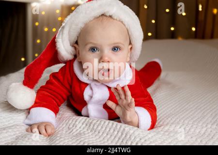 Schönes kleines Kind feiert Weihnachten. Neujahr. Ein Kind in einem Weihnachtskostüm. Neugeborenes Baby in Weihnachtsmann Hut über Lichter Hintergrund