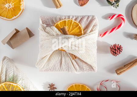 Weihnachtsgeschenk ohne Verschwendung. Hände, die Weihnachtsgeschenke in Leinenstoff mit Orangen dekorieren, grüner Tannenzweig auf weißem Tisch. Furoshiki-Geschenke. Stockfoto