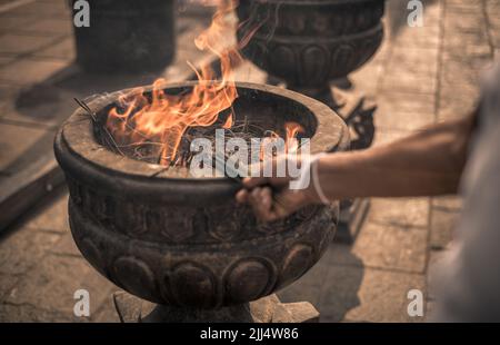 Neue Räucherstäbchen, die von der Hand der Frau angezündet werden, brennen Räucherstäbchen im alten Steintopf der Heiligen Sri Maha Bhodi Gärten. Stockfoto