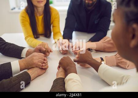 Ein vielfältiges Team von Menschen, die an einem Tisch sitzen und die Hände halten Stockfoto