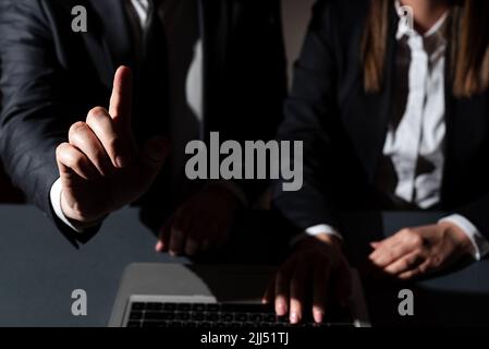 Sitzende Frau Auf Dem Schreibtisch Mit Einem Lap Top Und Einem Mann, Der Mit Einem Finger Auf Wichtige Botschaften Zeigt. Geschäftsmann Präsentiert Krutialinformationen Mit Einer Hand. Stockfoto