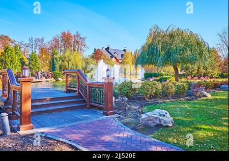 Die hölzerne Fußgängerbrücke auf dem See, umgeben von üppigem Grün, Weidenbäumen, gelben Birken und roten Eichen im Park, Mezhyhirya, Ukraine Stockfoto