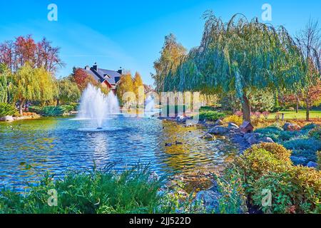 Idyllischer Herbstpark mit üppigen Weiden, gelben Birken, roten Eichen, Wacholderdickicht und See mit Brunnen, Mezhyhirya, Ukraine Stockfoto