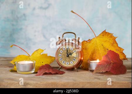 Wechsel von Sommer- auf Winterzeit, Vintage-Wecker mit einstündiger Rückstellung, Herbstblätter und Kerzen auf rustikalem Holz, hellblauer Hintergrund Stockfoto