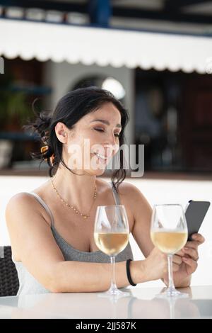 Erwachsene Frau, die in einem Restaurant im Freien sitzt und ihr Handy mit zwei Gläsern Wein auf dem Tisch benutzt Stockfoto