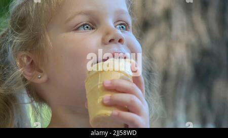 Nettes kleines Mädchen isst draußen Eis. Nahaufnahme eines blonden Mädchens, das auf der Parkbank sitzt und Eis isst. Stockfoto