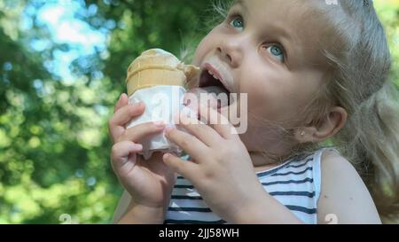 Nettes kleines Mädchen isst draußen Eis. Nahaufnahme eines blonden Mädchens, das auf der Parkbank sitzt und Eis isst. Stockfoto