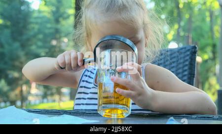 Nettes kleines Mädchen schaut vorsichtig in die Linse auf den Saft im Glas. Nahaufnahme eines blonden Mädchens, das ihren Saft im Glas studiert, während es sich ihr ansieht Stockfoto