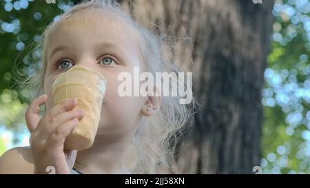 Nettes kleines Mädchen isst draußen Eis. Nahaufnahme eines blonden Mädchens, das auf der Parkbank sitzt und Eis isst. Stockfoto