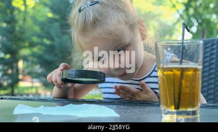 Kleines Mädchen schaut vorsichtig in die Linse auf das Salz. Nahaufnahme des blonden Mädchens studiert Salzkristalle, während sie durch die Lupe betrachtet wird Stockfoto