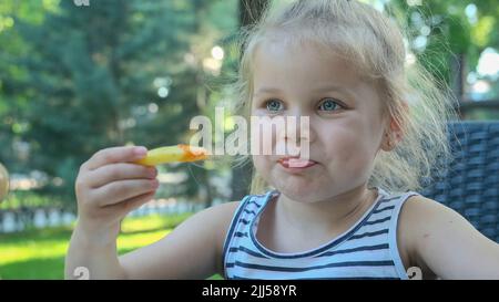 Kleine Mädchen essen pommes. Nahaufnahme des blonden Mädchens nimmt Kartoffelchips mit ihren Händen und probiert sie im Straßencafé im Park. Stockfoto