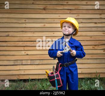 Niedlicher neugieriger Junge in blauen Overalls und einem Bauhelm mit Reparaturwerkzeugen in den Händen. Sei ein Baumeister wie Papa. Kleiner Helfer beim Erlernen neuer skil Stockfoto