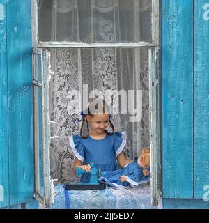 Porträt eines Schneidermädchen Nähen Sie Puppenkleidung auf einer Kinder-Nähmaschine im Fenster eines alten Holzhauses. Stockfoto