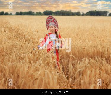 Kleines Mädchen im russischen nationalen Sarafan und ein Kokoschnik, der am Sommertag in einem goldenen Weizenfeld steht Stockfoto