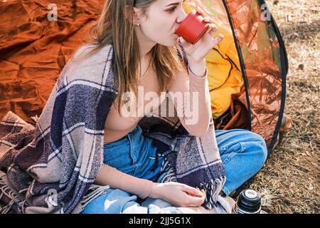 Glückliches junges Mädchen, das im Zelt sitzt und heißen Tee aus einer Tasse Thermoskanne vor dem Hintergrund des Frühlings- oder Herbstwaldes trinkt. Psychische Gesundheit. Zeit für sich selbst. Stockfoto