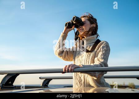 Glückliche junge Frau in warmer Kleidung, die durch ein Fernglas schaut Stockfoto