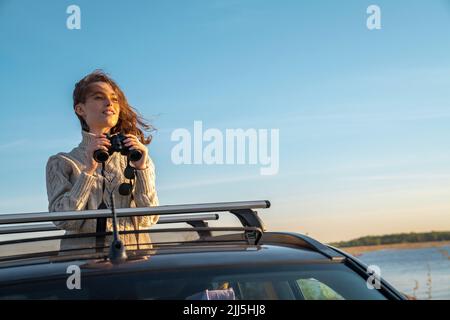 Lächelnde junge Frau mit einem Fernglas, das durch das Sonnendach des Autos steht Stockfoto
