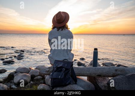 Junge Frau mit Rucksack, die bei Sonnenuntergang auf Holz sitzt Stockfoto