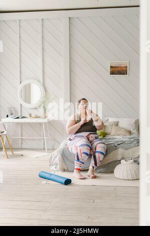 Frau, die zu Hause auf dem Bett saß und Wasser aus der Flasche trank Stockfoto