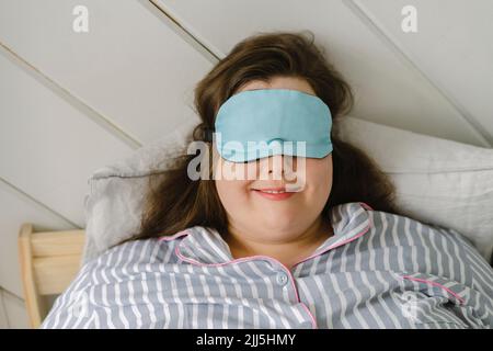 Lächelnde Frau mit schlafender Augenmaske, die zu Hause auf dem Bett liegt Stockfoto