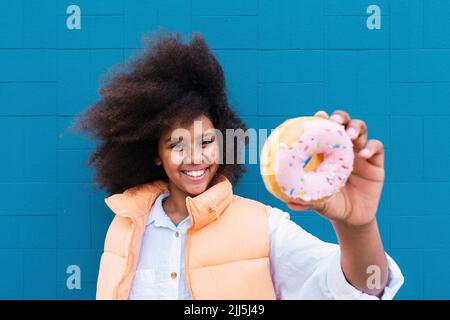 Lächelndes Mädchen, das Donut vor der blauen Wand zeigt Stockfoto