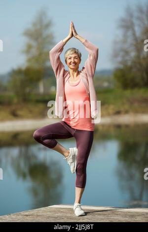 Glückliche reife Frau, die auf dem Pier die Baumhaltung übt Stockfoto