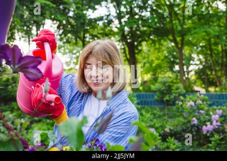 Lächelnde Frau, die Wasser auf blühende Pflanzen im Garten gießt Stockfoto