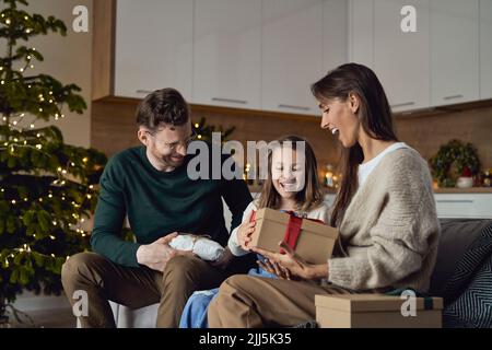 Glückliche Eltern schenken der Tochter, die auf dem Sofa sitzt, ein Weihnachtsgeschenk Stockfoto