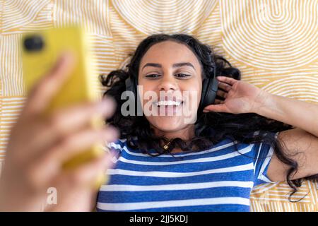 Glückliche Frau mit Kopfhörern und Smartphone im Schlafzimmer Stockfoto