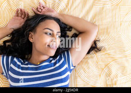 Nachdenkliche Frau mit erhobenen Armen, die zu Hause auf dem Bett lag Stockfoto