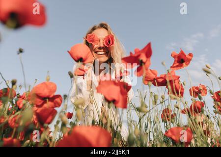 Glückliche Frau, die im Mohnfeld die Augen mit Blumen bedeckt Stockfoto