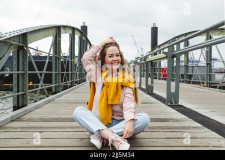 Glückliche junge Frau mit der Hand im Haar, die auf dem Steg sitzt Stockfoto